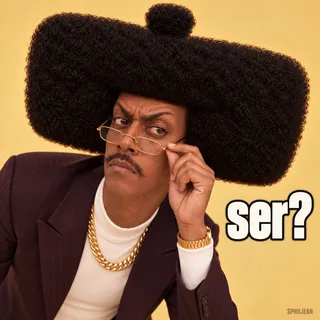 Phil leaning close toward the camera over tiny gold reading glasses, one eyebrow raised in extreme polite disbelief, his full huge rectangular afro and long neck visible, warm plain cream background.