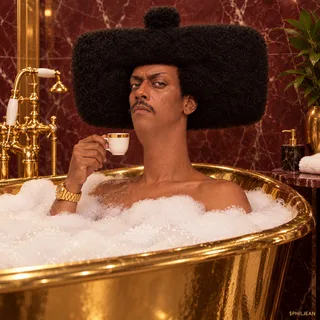 Phil sitting in a freestanding gold bathtub filled with white bubbles, only head shoulders and one hand with gold watch visible, holding a tiny espresso cup, burgundy marble bathroom, entirely deadpan, nonsexual.