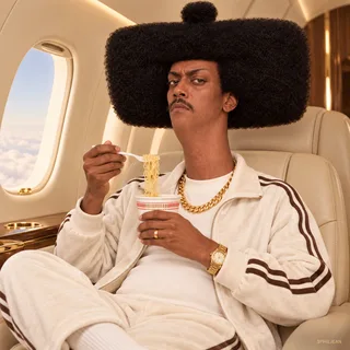 Phil in cream designer tracksuit and gold chain sitting alone in a beige private jet, casually eating cheap instant noodles with a tiny plastic fork, unimpressed stare, full afro intact and clearly visible, flash editorial.