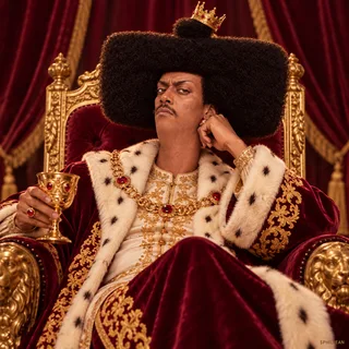 Phil on an extravagant burgundy velvet throne in gold embroidered robe, tiny crooked gold crown perched on the small top nub of his giant rectangular afro, exhausted king energy, full hair silhouette, theatrical portrait.