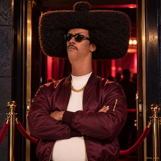 Phil outside a velvet-rope nightclub in burgundy bomber jacket, black sunglasses and gold chain, arms crossed as a stern bouncer, he alone occupies practically the entire doorway with his hair, dramatic night flash.